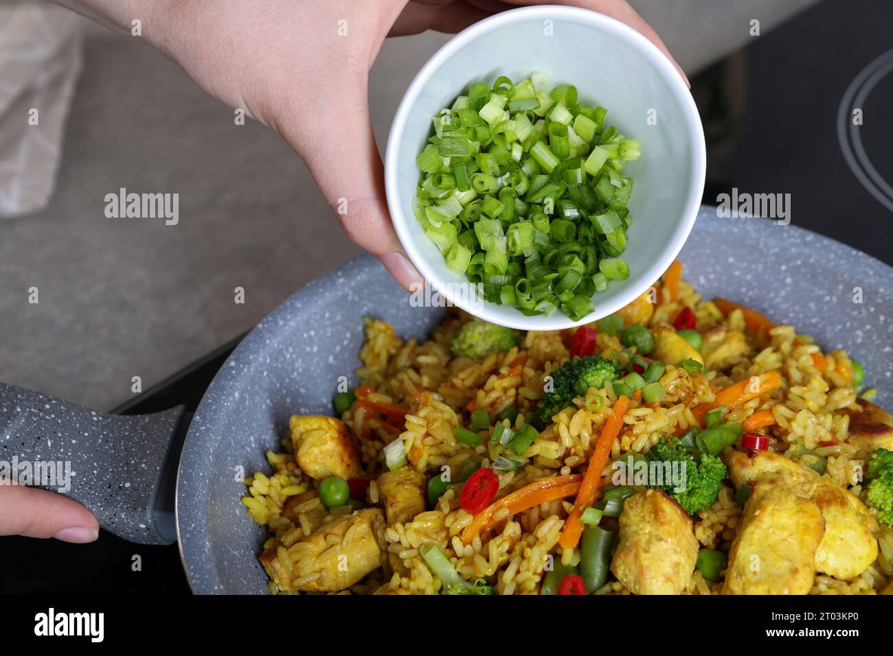 Frau fügt geschnittene grüne Zwiebeln zu Reis mit Fleisch und Gemüse in der Pfanne hinzu, Nahaufnahme Stockfoto