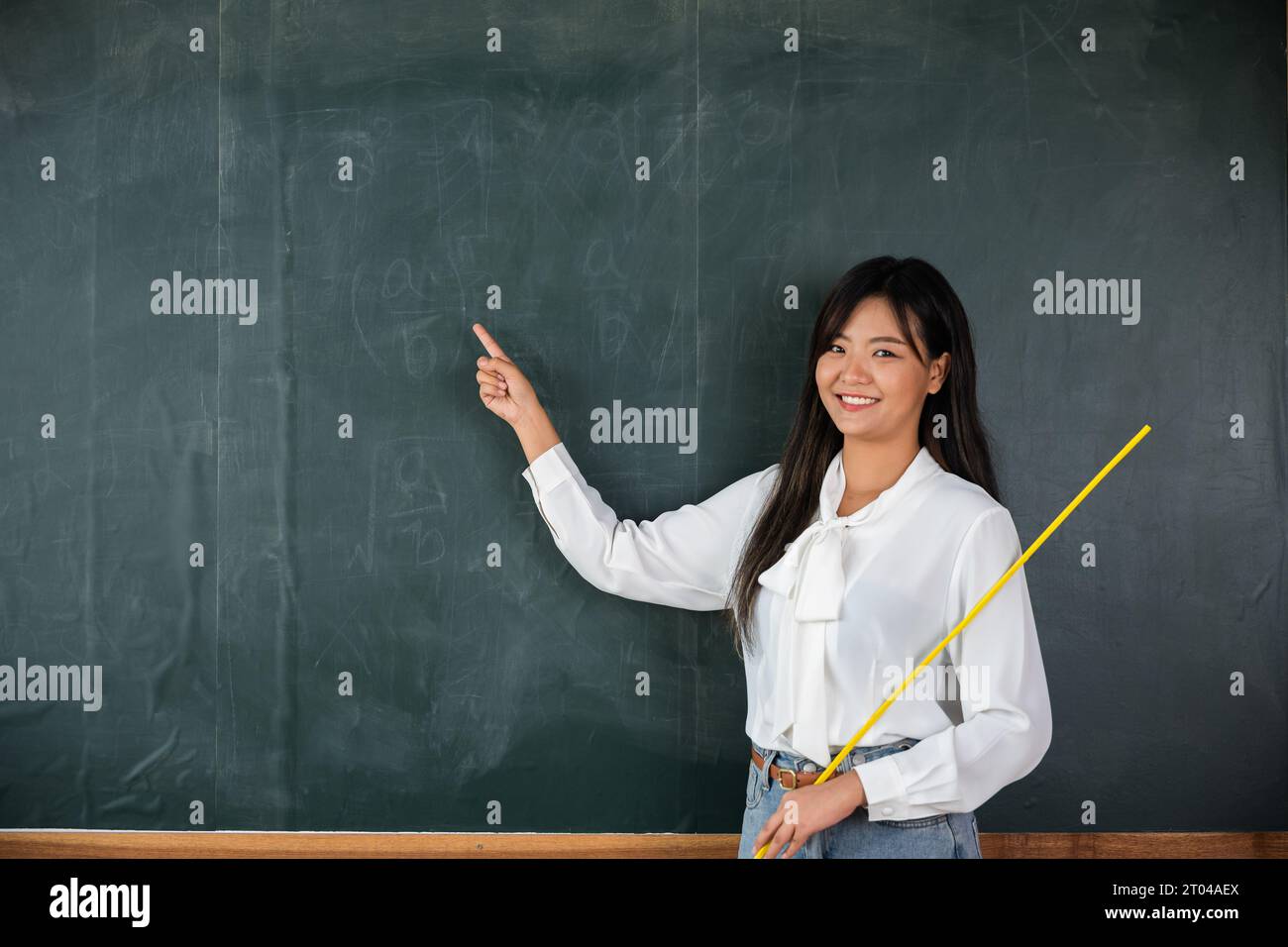 Asiatische Lehrerin lächelt mit Holzstab, der auf die Tafel zeigt Stockfoto