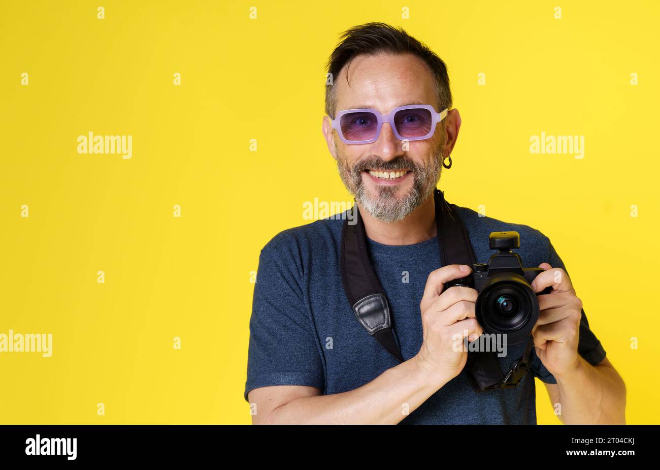 Der glückliche, begeisterte Fotograf lächelt vor Freude, während er die spiegellose Kamera vor gelbem Hintergrund hält. Freude und Leidenschaft, die Fotografen spüren, wenn sie Momente mit der geliebten Kamera festhalten. Hochwertige Fotos Stockfoto