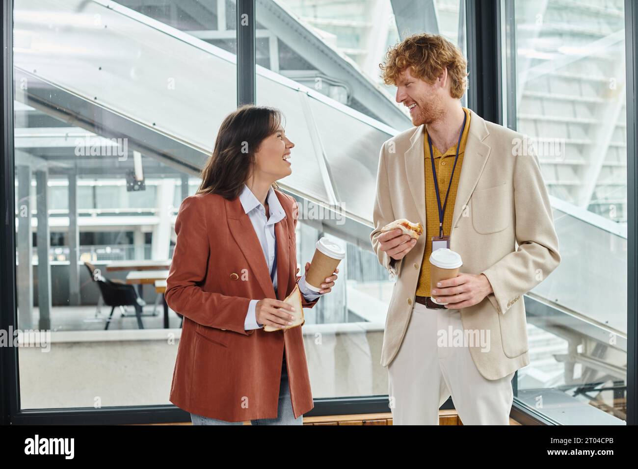 Junge Kollegen genießen Mittagspause und lächeln einander mit Glas vor dem Hintergrund an, arbeiten zusammen Stockfoto