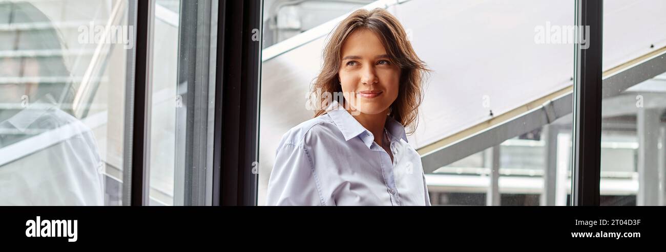Fröhliche junge Frau in elegantem Outfit, die wegblickt mit Fensterkulisse, Coworking-Konzept, Banner Stockfoto