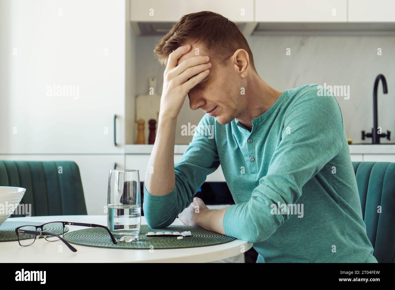 Mann in türkisfarbenem T-Shirt sitzt zu Hause am Küchentisch und hält seinen Kopf mit der Hand. Krankheit und Einsamkeit. Auf dem Tisch steht ein Glas Wasser und Stockfoto