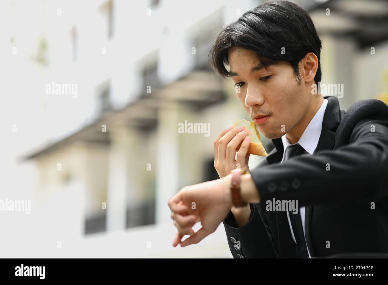 Beschäftigter asiatischer Büroangestellter in Eile, der Sandwiches beißt und die Zeit auf der Armbanduhr für einen Termin oder Transport überprüft Stockfoto