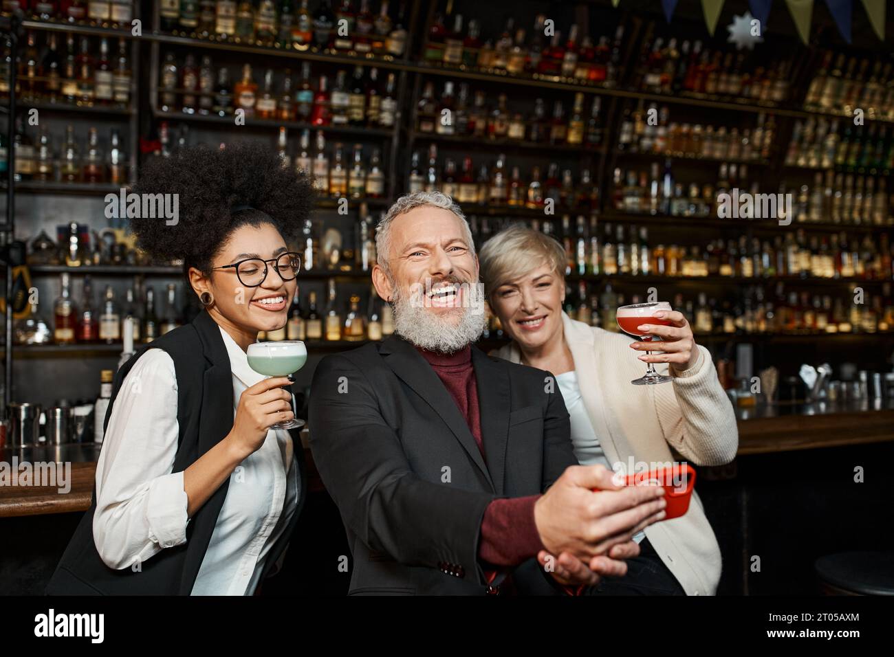 Fröhlicher bärtiger Mann, der Fotos mit multiethnischen Frauen in der Bar macht, ein vielfältiges Team, das sich nach der Arbeit ausruhen lässt Stockfoto