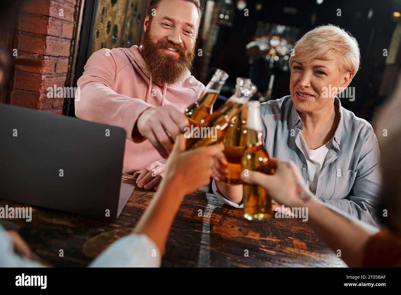 Fröhlicher bärtiger Mann und Frau mittleren Alters, die Bierflaschen mit Kollegen in der Nähe des Laptops im Pub anklickt Stockfoto