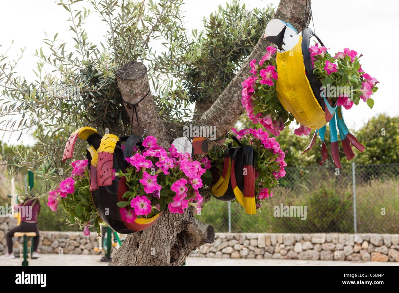 Bunte Papageiendekorationen, die am Baum hängen, mit Autoreifen und rosa Petunien auf „Costitx en Flor“ (Costitx in Blüte), Blumenmesse, Mallorca, Stockfoto