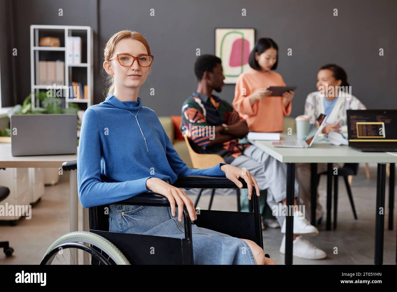 Porträt einer jungen Frau im Rollstuhl mit Blick auf die Kamera in einem modernen Büro mit Team im Hintergrund, Kopierraum Stockfoto