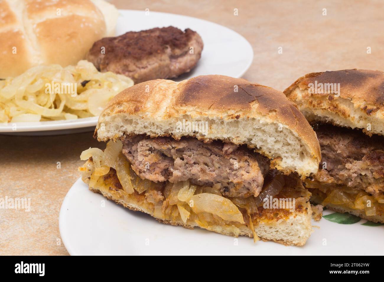 Eine Nahaufnahme eines hausgemachten Hamburger, der in zwei Hälften geschnitten ist und auf frisch gekochten Zwiebeln zwischen gerösteten Burgerbrötchen liegt. Im Hintergrund ist da Stockfoto
