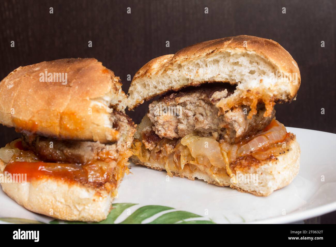 Köstlicher hausgemachter Hamburger mit Zwiebeln und Tomaten auf rustikalem Brot, auf einem weißen Teller vor dunklem Holzhintergrund. Perfekter Gourmet in der Nähe Stockfoto