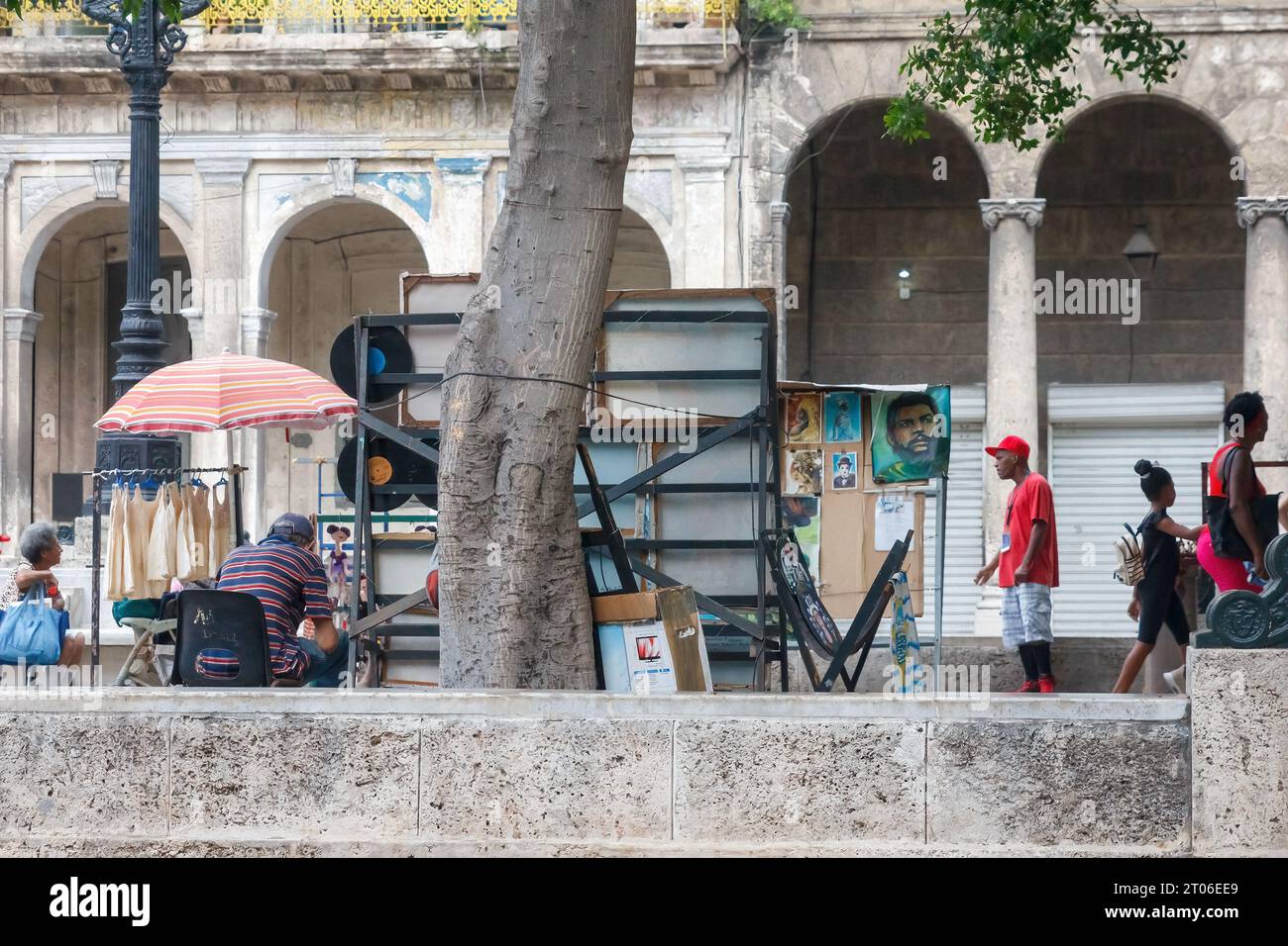 Ein Mann verkauft Kunst in Paseo del Prado. Er sitzt unter einem Sonnenschirm. Andere Kubaner laufen tagsüber durch die Szene. Stockfoto