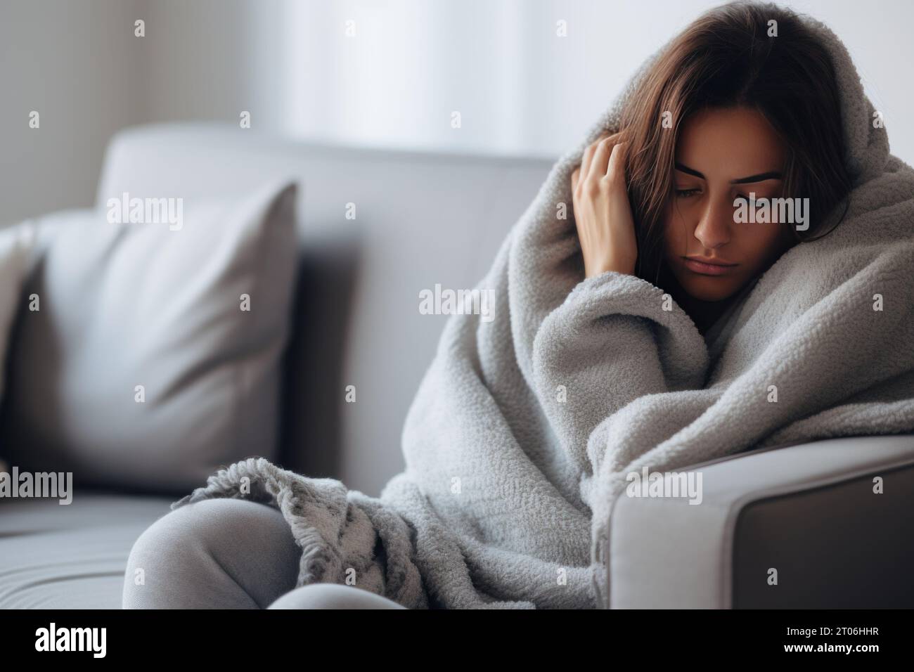 Eine Frau auf ihrem Heimsofa, in eine Decke gewickelt, vor der Kälte zitternd und einen besorgten Ausdruck mit Copyspace trägt Stockfoto