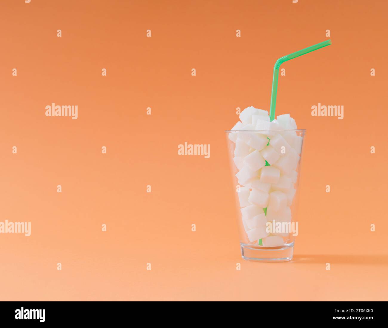 Kreative Komposition aus Glas voller Zuckerwürfel mit grünem Trinkstroh auf pastellorangenem Hintergrund. Minimales ungesundes Ernährungskonzept. Stockfoto