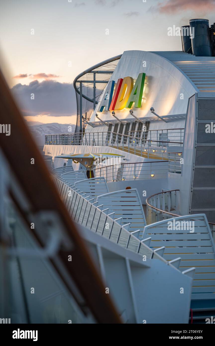 Beleuchtetes AIDA-Logo auf dem Kreuzfahrtschiff AIDA Bella bei Sonnenuntergang, Zaun der Treppe und Bootsdeck vorne, vertikale Aufnahme Stockfoto