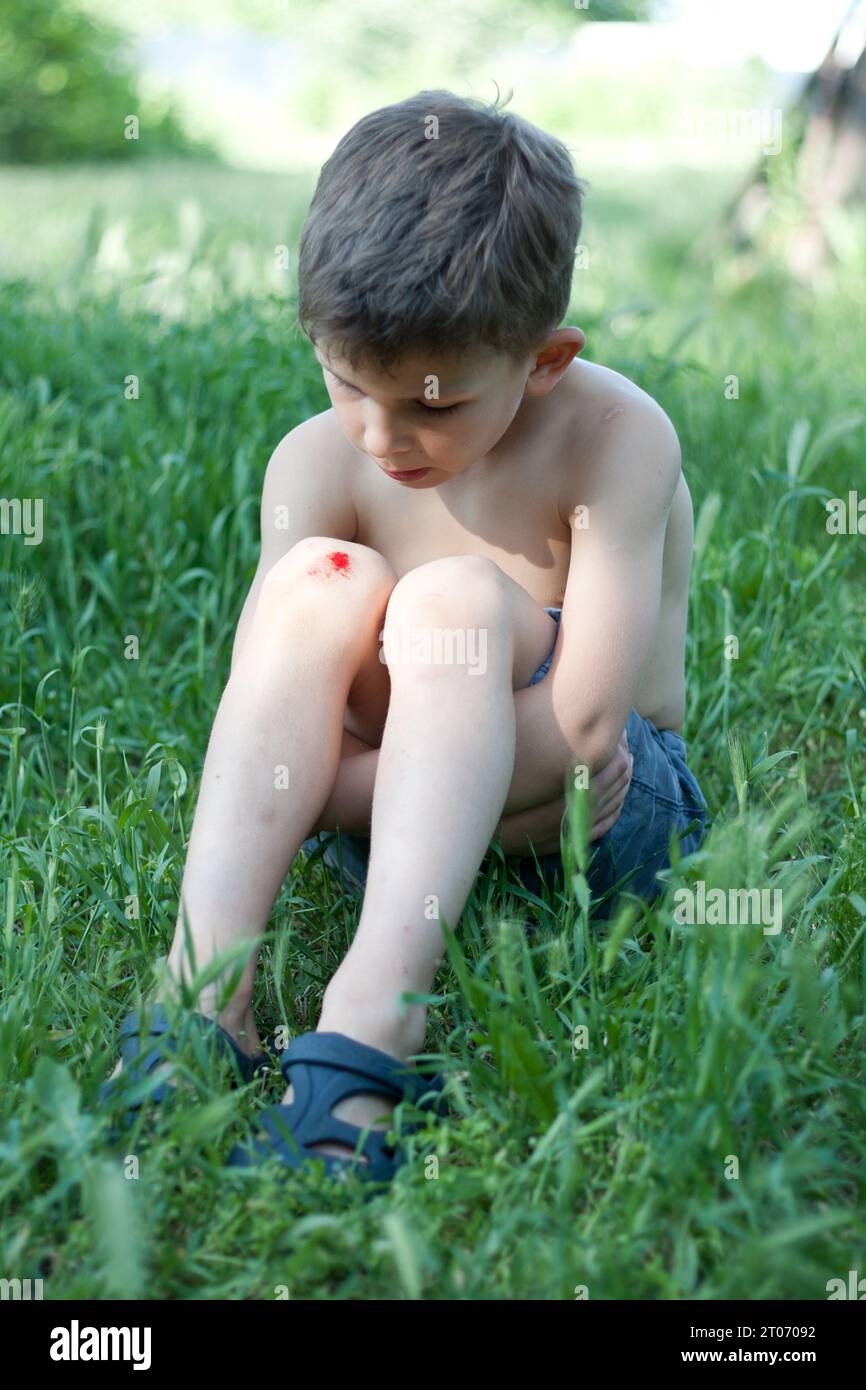Porträt eines auf Gras sitzenden Vorschuljungen mit frischer Blutung am Knie. Kind fiel, zerkratzte Haut am Knie, verletzt, verärgert. Kinderverletzungen bei su Stockfoto