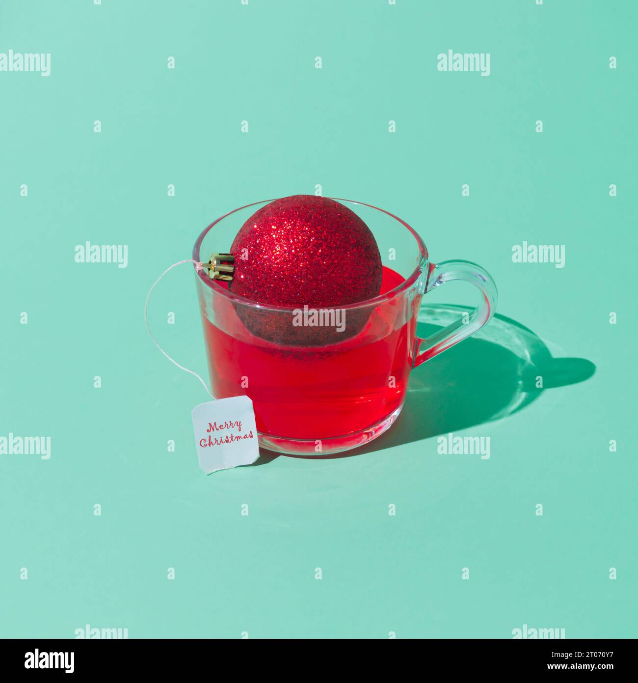 Kreative Weihnachtskomposition aus transparentem Glasbecher Fruchttee mit Teebeutel aus roter Weihnachtskugel auf grünem Hintergrund. Minimales Konzept. Stockfoto