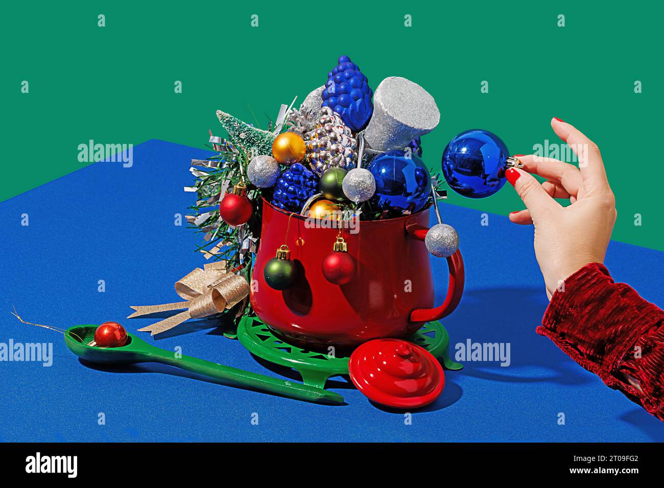 Beschneiden Sie anonymes Weibchen mit roten Nägeln, die weihnachtskugel in einem Topf mit verschiedenen Ornamenten nahe dem Löffel auf blauem und grünem Hintergrund platzieren Stockfoto