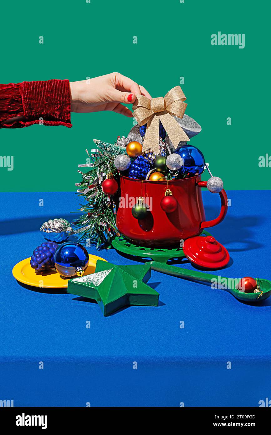 Beschneiden Sie anonymes Weibchen mit roten Nägeln, die weihnachtsbandkegel in einen Topf mit verschiedenen Ornamenten neben Teller und Löffel gegen blaues und grünes Backgro legen Stockfoto