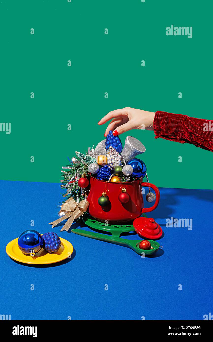Beschneiden Sie anonymes Weibchen mit roten Nägeln und legen Sie weihnachts-Ananaskegel in einen Topf mit verschiedenen Ornamenten in der Nähe von Teller und Löffel auf blauem und grünem Rücken Stockfoto