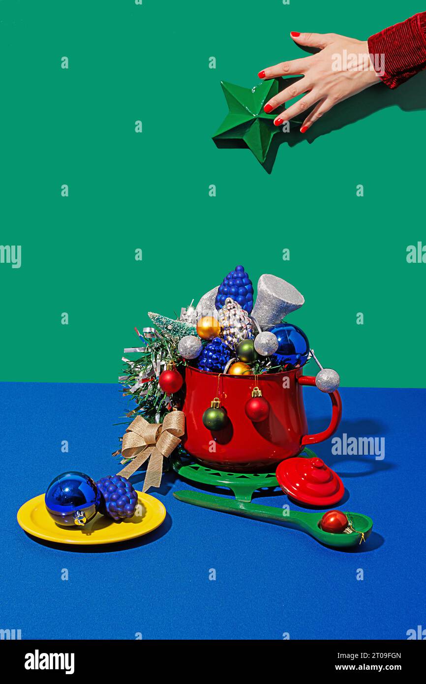 Ernten Sie anonymes Weibchen mit roten Nägeln, die einen grünen weihnachtsstern über dem Topf halten, mit verschiedenen Ornamenten in der Nähe des Tellers und des Löffels gegen blauen und grünen Hintern Stockfoto