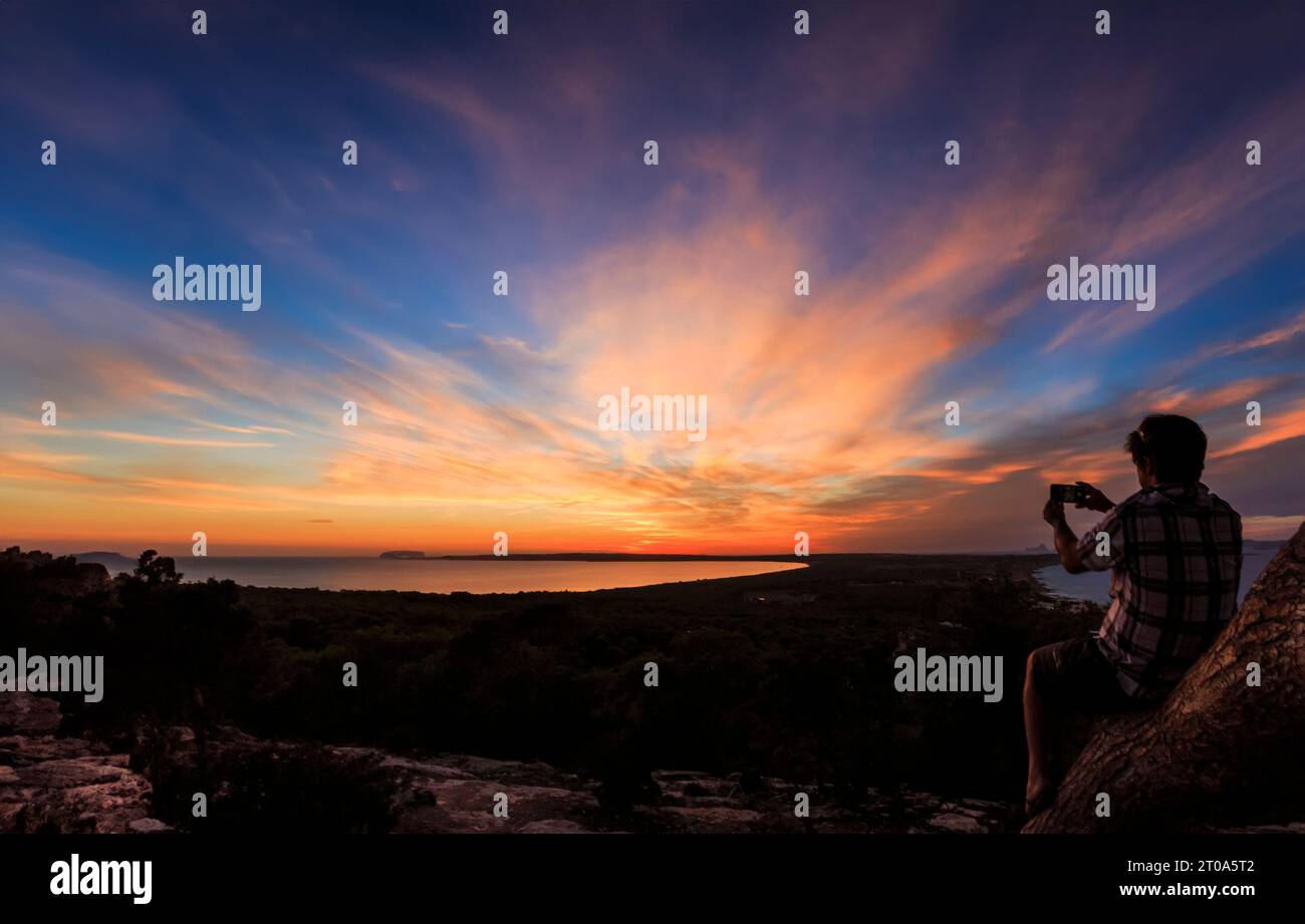 An Formenteras ruhigen Ufern sitzt ein Mann auf einem Baumstamm und nimmt mit seinem Handy den faszinierenden Sonnenuntergang auf. Die goldenen Töne des Horizonts ju Stockfoto