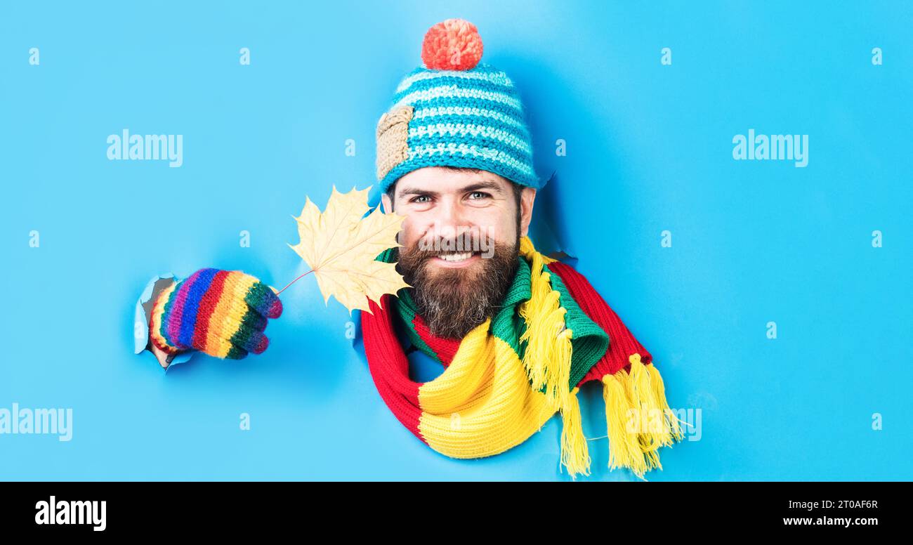 Lächelnder bärtiger Mann in modischem bunten Strickmütze, Schal und Handschuhen, der durch das Papierloch schaut. Schöner Kerl in warmen Kleidern mit Ahornblatt Stockfoto