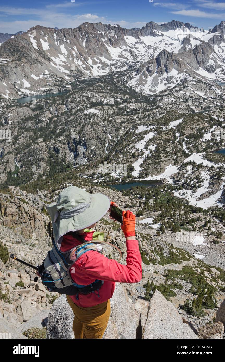 Eine Tageswandererin mit großem Sonnenhut macht ein Handyfoto vom Bergrücken der Sierra Nevada mit Blick auf das Becken des Lake Sabrina Stockfoto