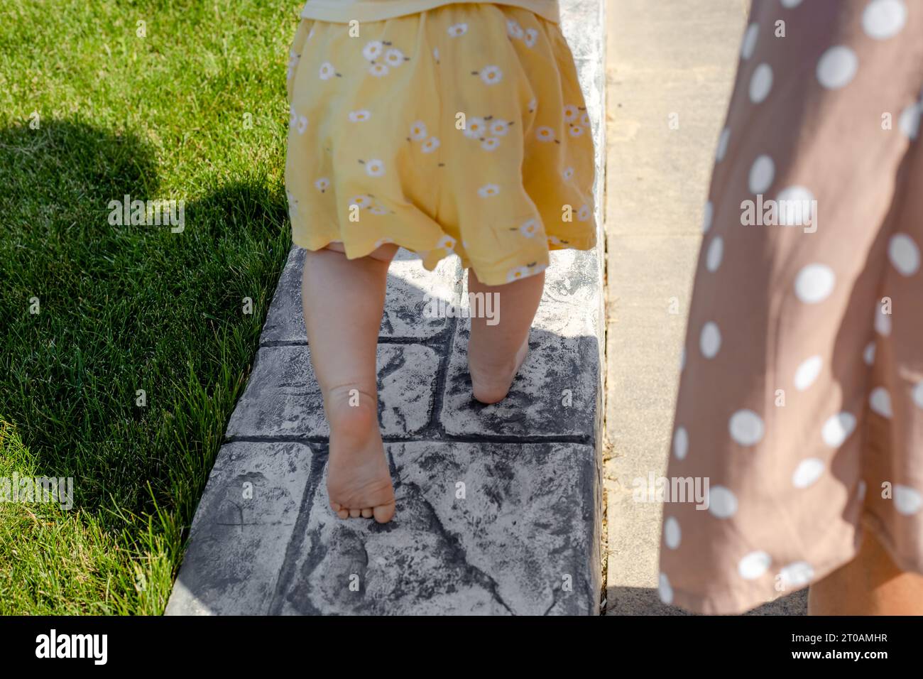Ein kleines Kind läuft barfuß auf dem Rasen entlang der Bordsteinkante, Rückansicht. Die ersten Schritte Ihres Babys. Stockfoto