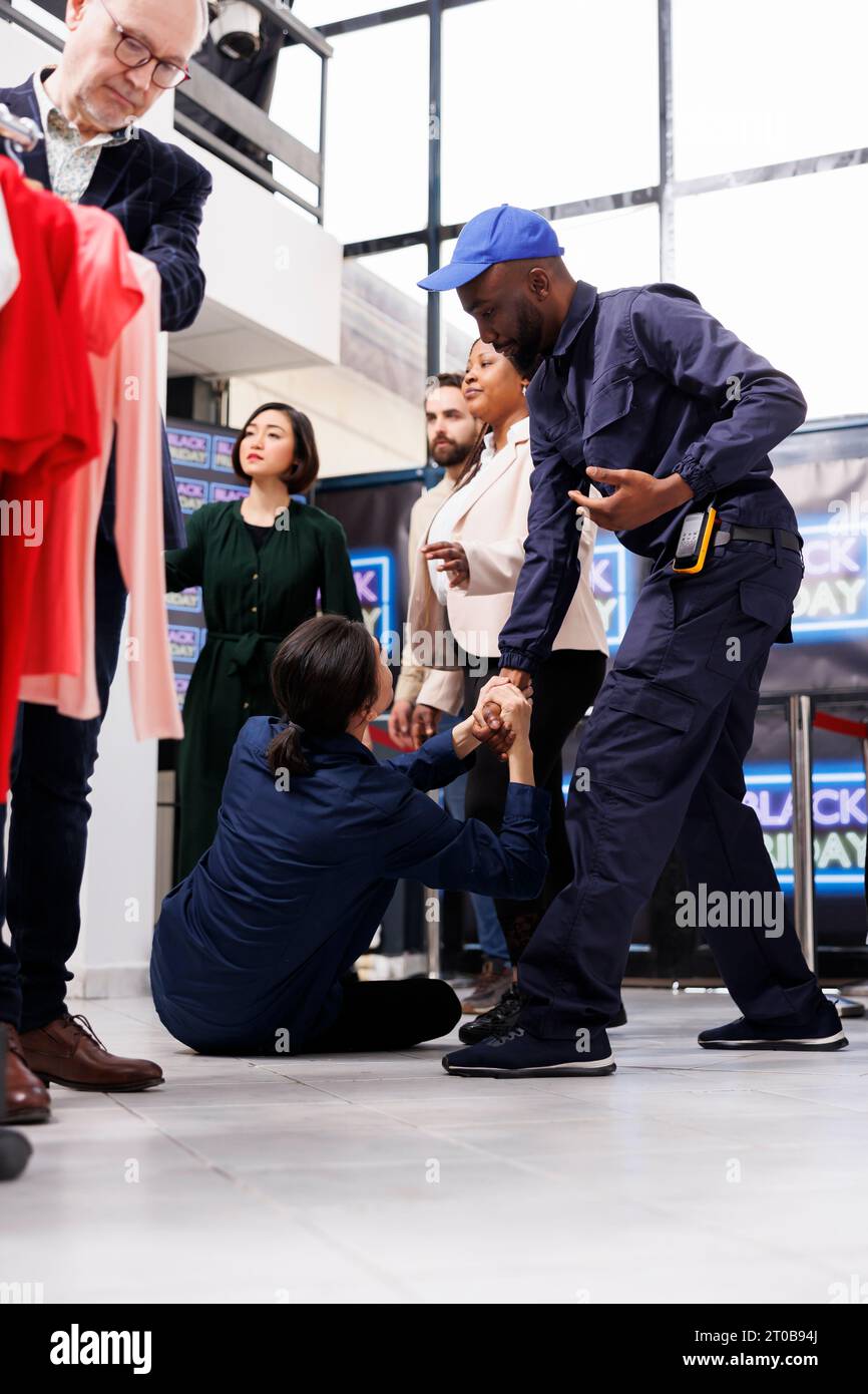 Black Friday Shopper verletzt. Der Sicherheitsbeamte des afroamerikanischen Mannes trägt Uniform, um die Sicherheit von Bekleidungsgeschäften und Kunden während der hektischen Verkaufssaison zu gewährleisten und den Kunden dabei zu helfen, vom Boden aufzustehen Stockfoto