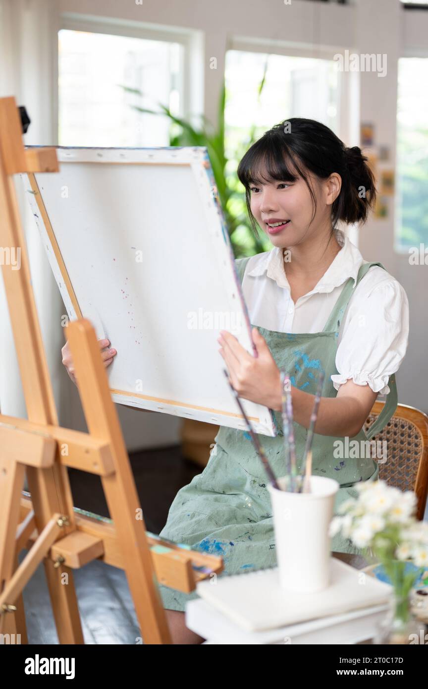 Eine zufriedene und charmante junge asiatische Künstlerin in einer Schürze mit Leinwand, die ihr gemaltes Bild in ihrem minimalistischen Atelier ansieht. Stockfoto