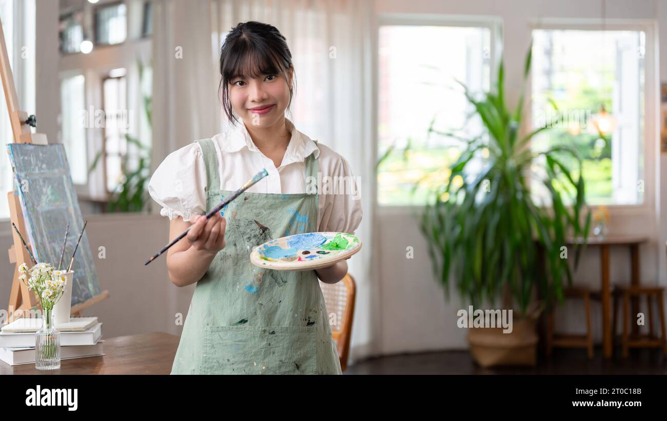 Ein Porträt einer charmanten und lächelnden jungen asiatischen Künstlerin in einer Schürze mit Pinsel und Farbpalette, die in ihrem Atelier steht. Stockfoto