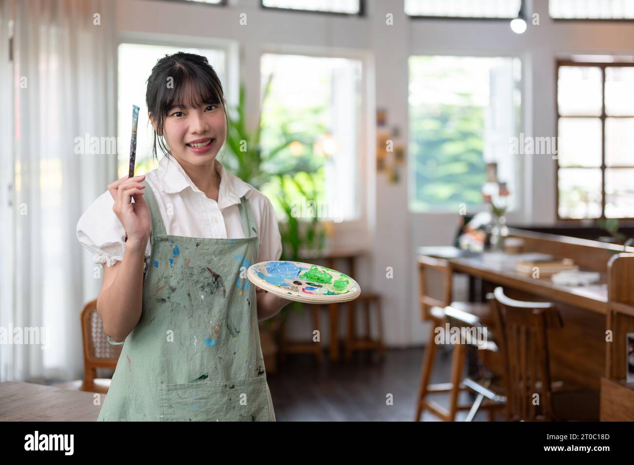 Ein Porträt einer charmanten und lächelnden jungen asiatischen Künstlerin in einer Schürze mit Pinsel und Farbpalette, die in ihrem Atelier steht. Cre Stockfoto