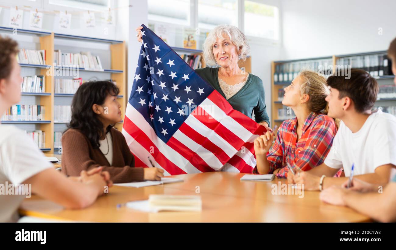 Reife Lehrerin, die mit Schulmädchen und Schuljungen arbeitet und die Nationalflagge der Vereinigten Staaten von Amerika in der Bibliothek hält Stockfoto