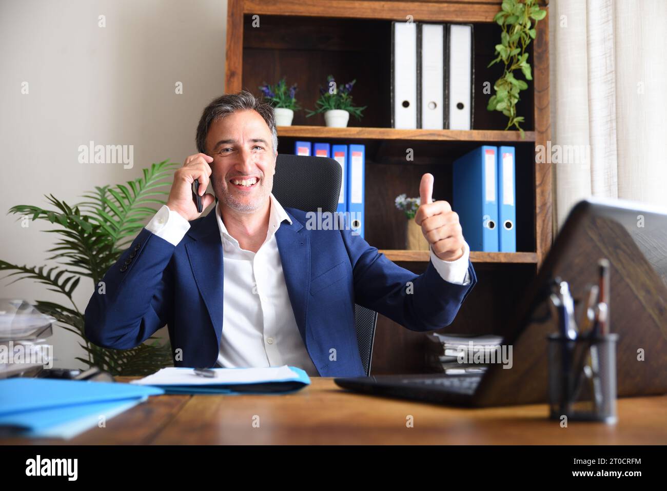 Ein lächelnder Geschäftsmann, der in seinem Büro auf dem Handy spricht, mit der Hand, die das OK-Schild mit Dokumenten und Laptop auf dem Tisch und Regalen zeigt Stockfoto