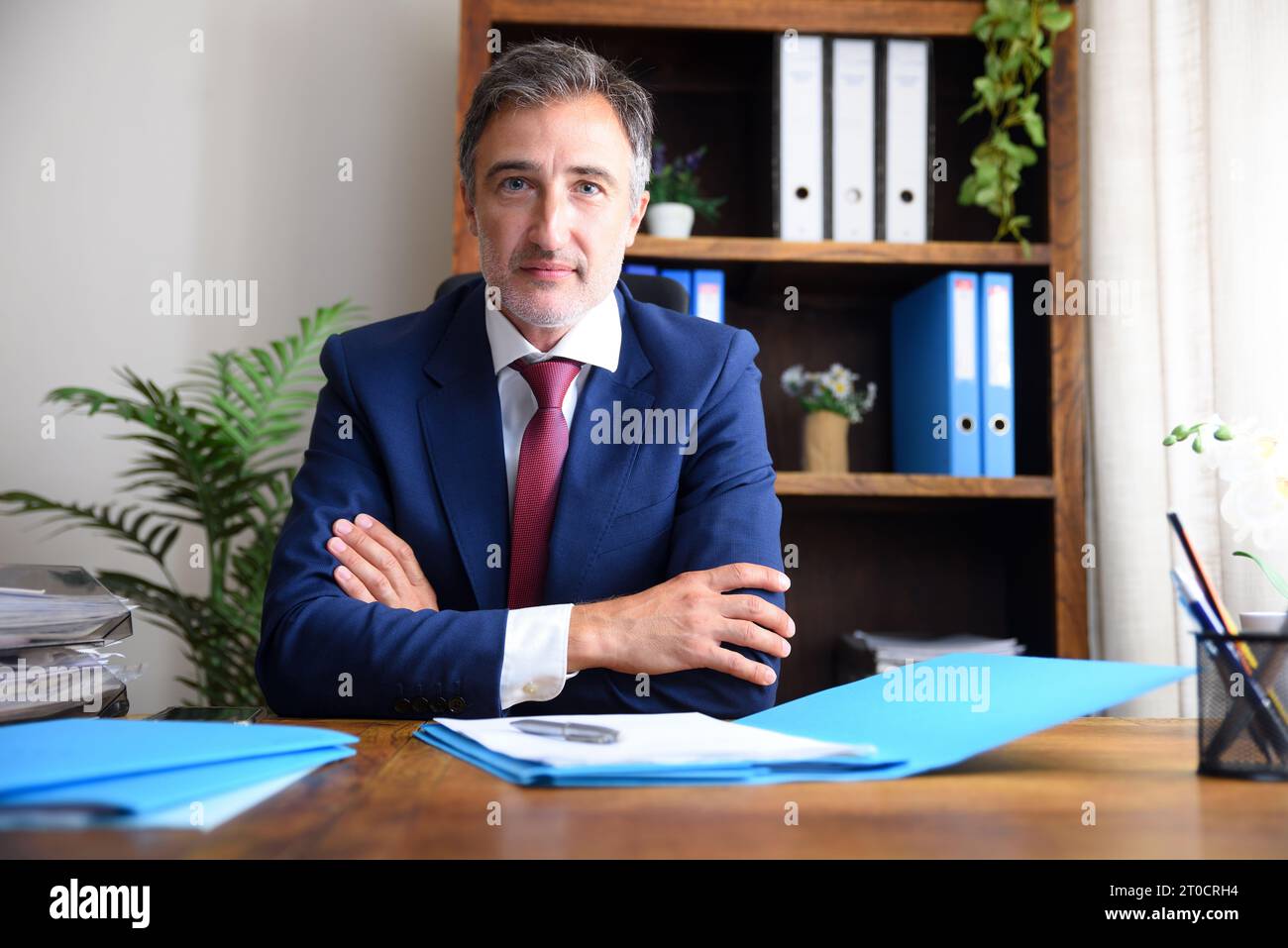 Geschäftsmann in seinem Heimbüro in blauem Anzug und roter Krawatte mit Dokumenten auf Holztisch und Bücherregalen Hintergrund. Stockfoto