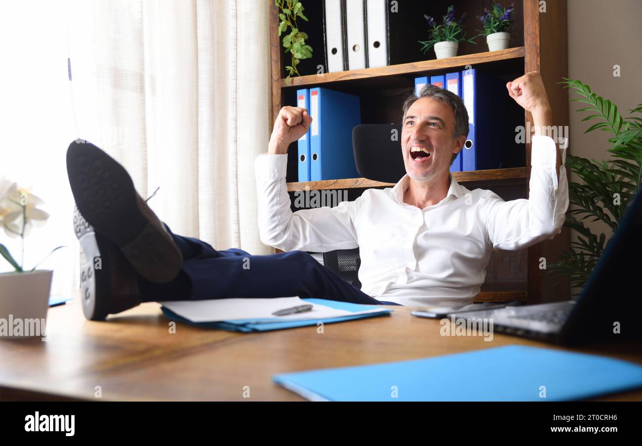 Euphorischer Geschäftsmann feiert Arbeitserfolg mit erhobenen Fäusten und Füßen auf dem Tisch in einem Büro mit Holzmöbeln Stockfoto