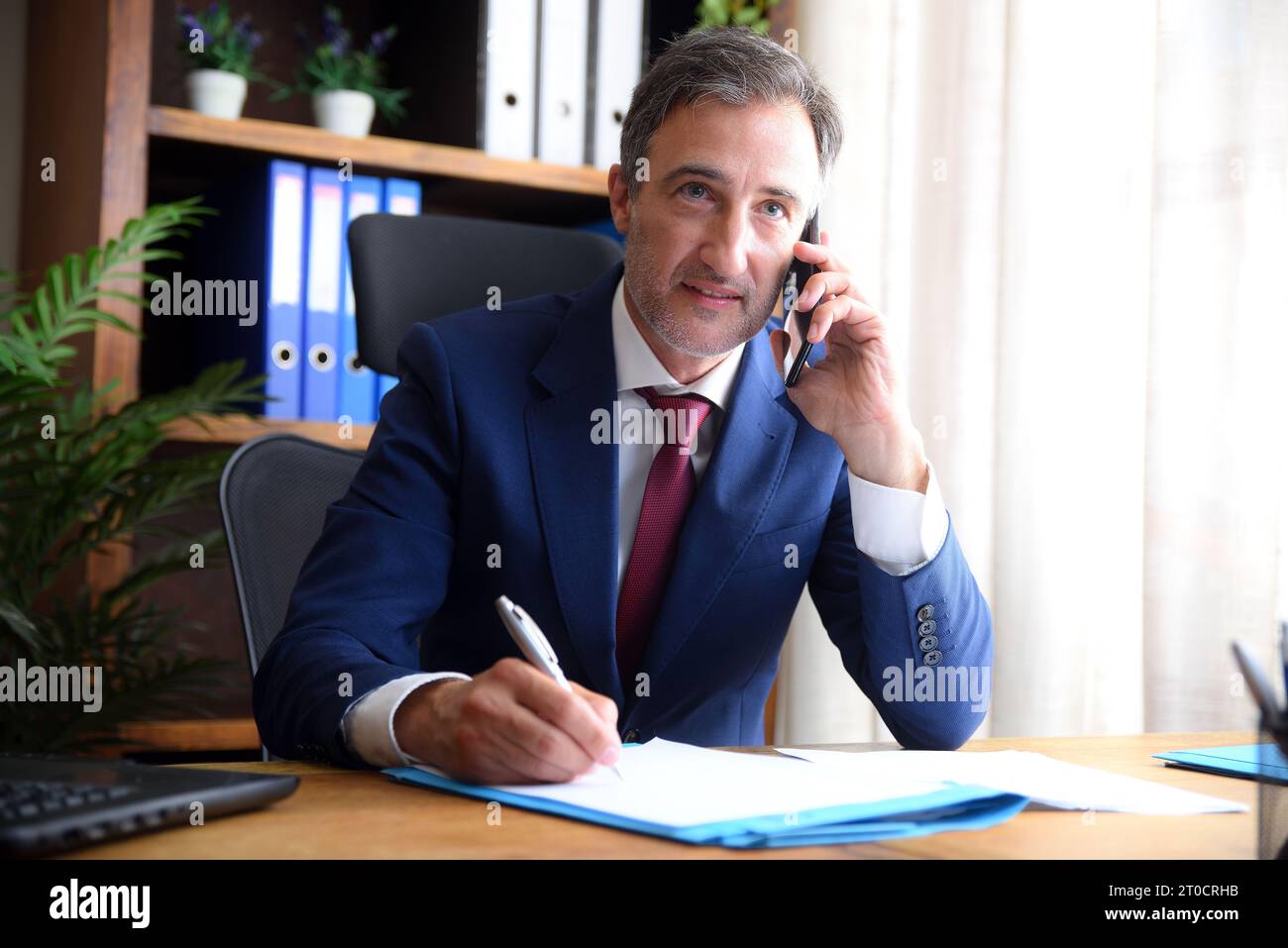 Geschäftsmann in blauem Anzug und Krawatte, der auf dem Handy spricht und im Büro schreibt, mit Dokumenten auf dem Tisch und Regalen im Hintergrund Stockfoto