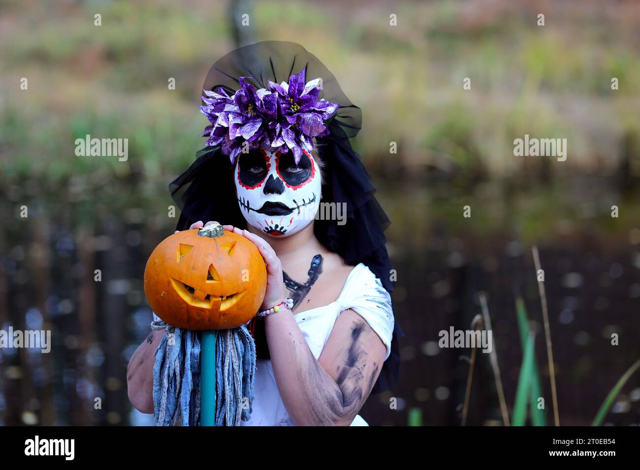 Ernsthaftes Mädchen im gruseligen Dead Bride Outfit mit Kürbis Jack o Laterne, die in die Kamera blickt, während sie während der Halloween-Feiertage in der Natur steht Stockfoto