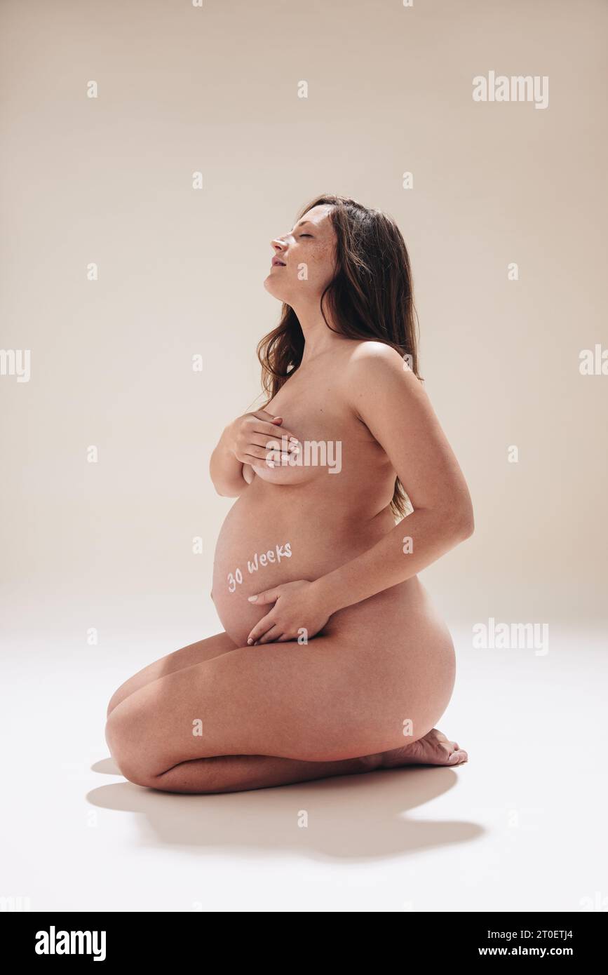 In ihrem dritten Trimenon umarmt eine selbstbewusste Kaukasierin ihren Babybump und strahlt ein positives Körperbild aus. Dieses Antennenporträt feiert den Beau Stockfoto