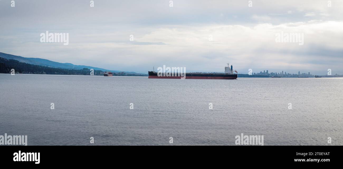 Frachtschiffe ankern am frühen Morgen außerhalb der Innenstadt. Panoramablick auf die Meereslandschaft mit Containerschiffen, Skyline der Stadt und Bergen. Selektiv Stockfoto