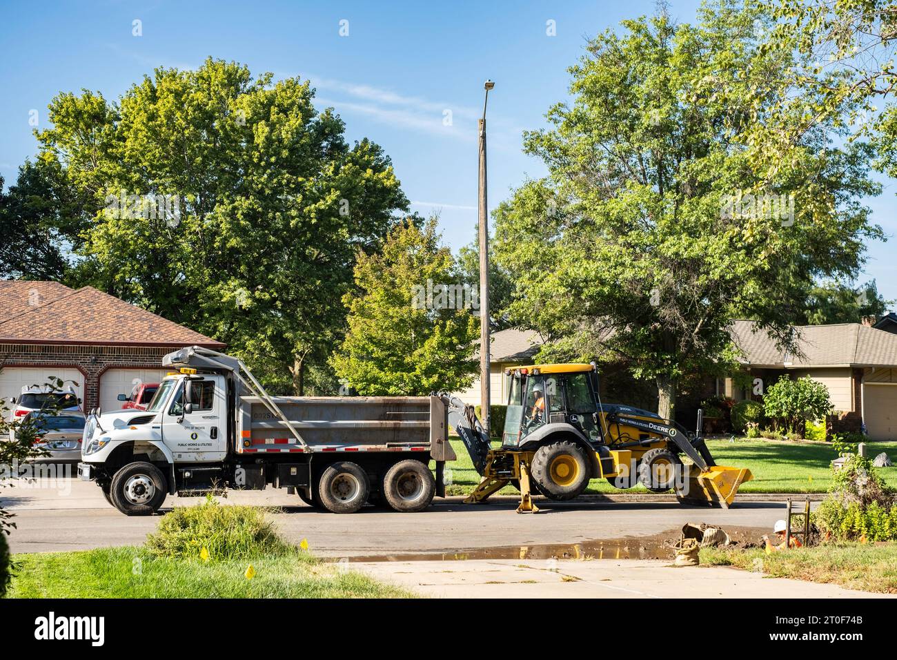 Ein Mitarbeiter der Wasserabteilung von Wichita Kansas gräbt eine Wasserleitung mit John Deere Baggerlader mit Bulldozer in einem Stadtviertel aus. Wichita, Kansas, USA. Stockfoto