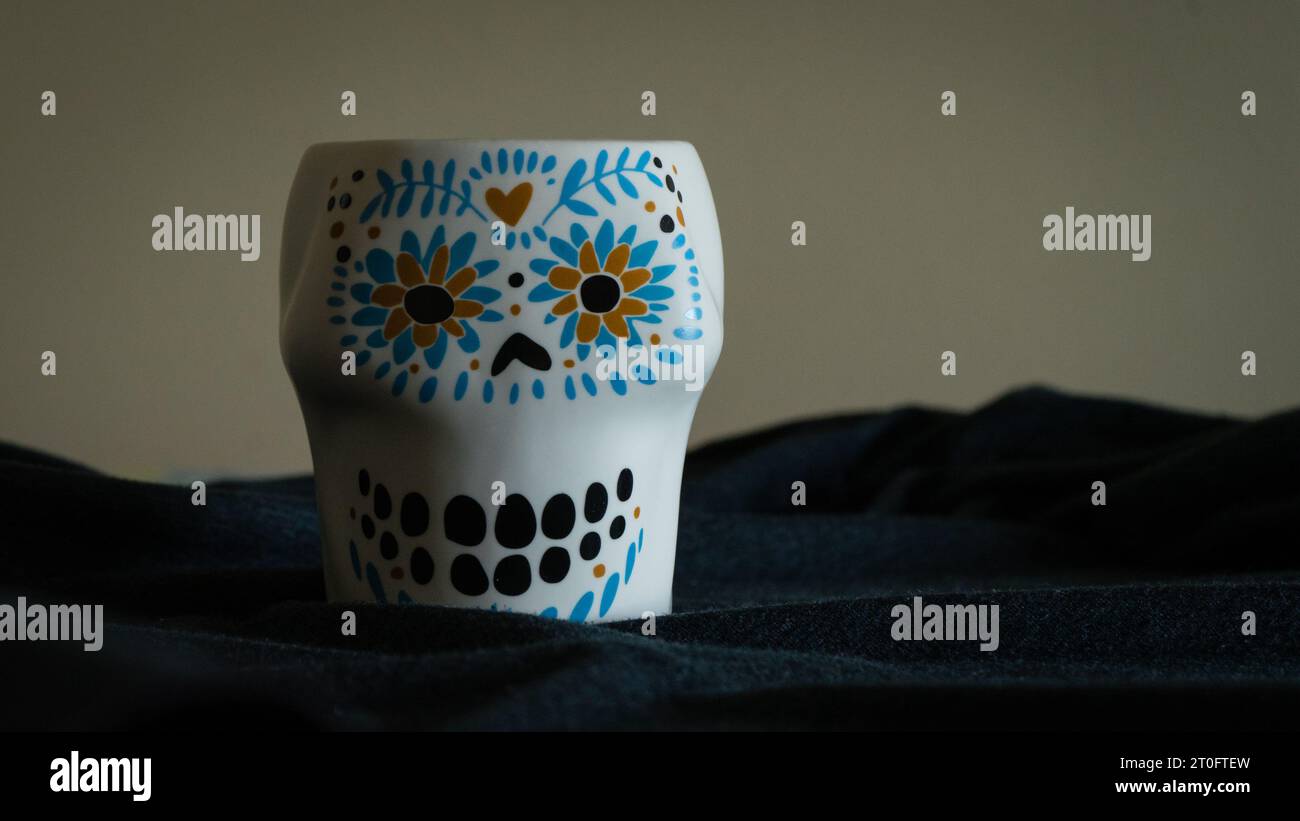Ein Becher mit Schädelgesicht setzt vor einem geheimnisvollen grau-schwarzen Hintergrund ein markantes Statement, das makabere Ästhetik mit moderner Raffinesse verbindet Stockfoto