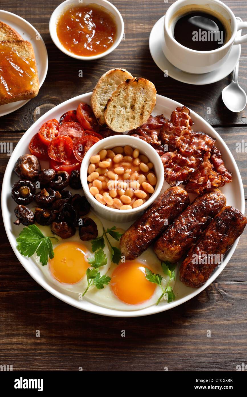 Komplettes englisches Frühstück mit Spiegelei, Speck, Würstchen, Bohnen und Pilzen auf einem Holztisch. Stockfoto