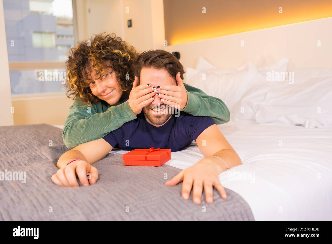 Valentinstag, ein junges kaukasisches Paar im Pyjama auf dem Bett, die Braut gibt eine nette Überraschung mit einem Geschenk an den Freund Stockfoto