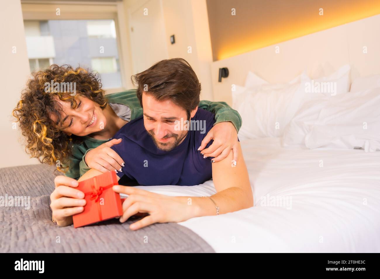 Valentinstag, an ein junges kaukasisches Paar im Pyjama auf dem Bett, die Braut gibt dem Freund eine nette Überraschung mit einem Geschenk. Mit Kopie und Stockfoto