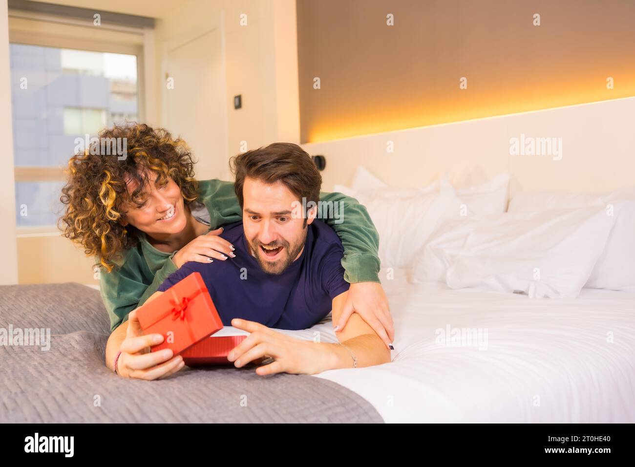 Valentinstag, an ein junges kaukasisches Paar im Pyjama auf dem Bett, die Braut gibt dem Freund eine nette Überraschung mit einem Geschenk. Mit Kopie und Stockfoto
