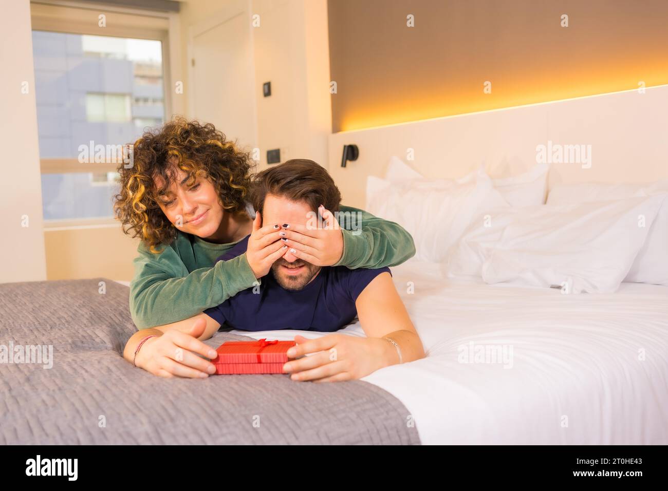 Valentinstag, an ein junges kaukasisches Paar im Pyjama auf dem Bett, die Braut gibt dem Freund eine nette Überraschung mit einem Geschenk. Mit Kopie und Stockfoto