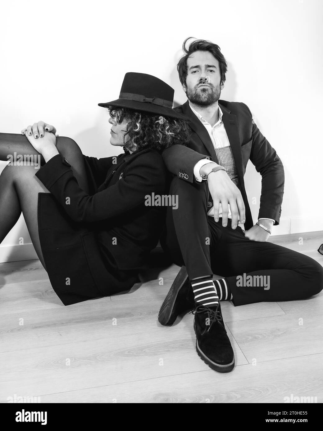 Ein kaukasisches Paar, das in einem schwarz-weißen Modeschuss auf dem Boden sitzt, in eleganten Anzügen und mit seriösem Aussehen Stockfoto