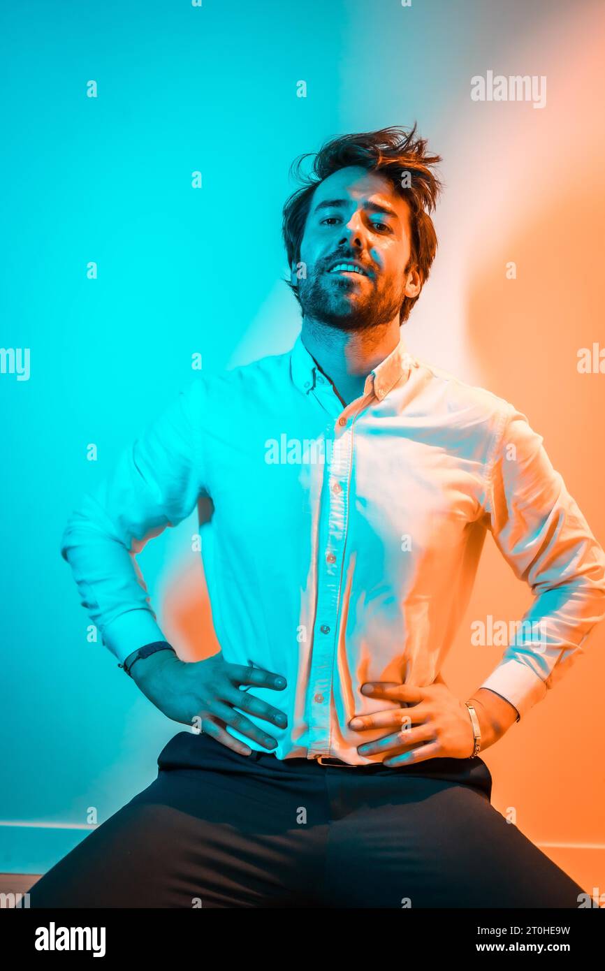 Studio Lifestyle, ein kaukasischer Mann auf den Knien in einem weißen Hemd und einer schwarzen Hose, beleuchtet von einem orange-blauen Neonlicht Stockfoto