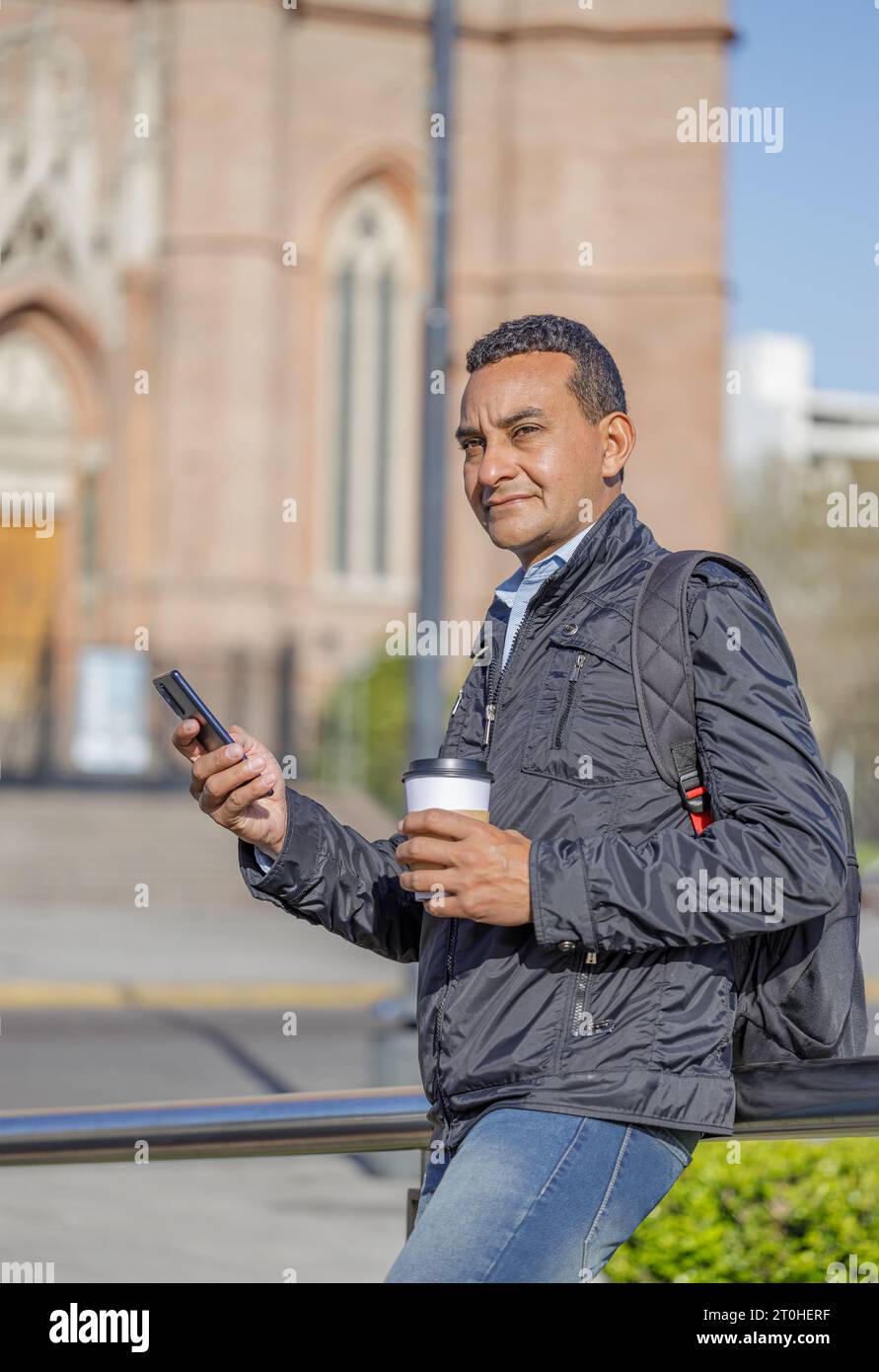 Porträt eines Latino-Mannes, der sein Handy benutzt und eine Tasse Kaffee hält, die auf einem Geländer in einem öffentlichen Park steht. Stockfoto