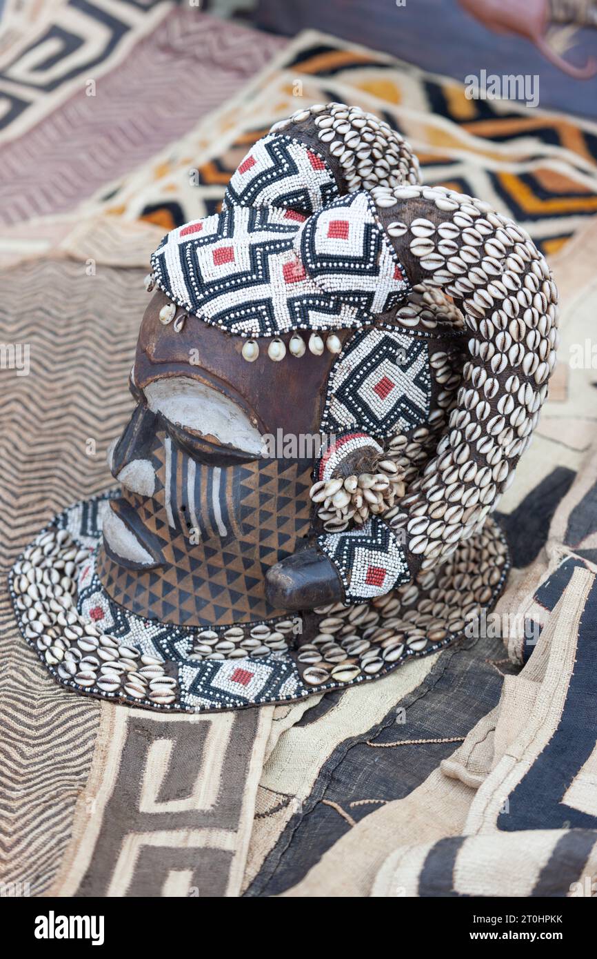 Vintage afrikanische Holzmaske mit Muscheln, die eine verwitterte Patina zeigen, auf traditionellen Teppichen bei einem Straßenverkäufer Stockfoto